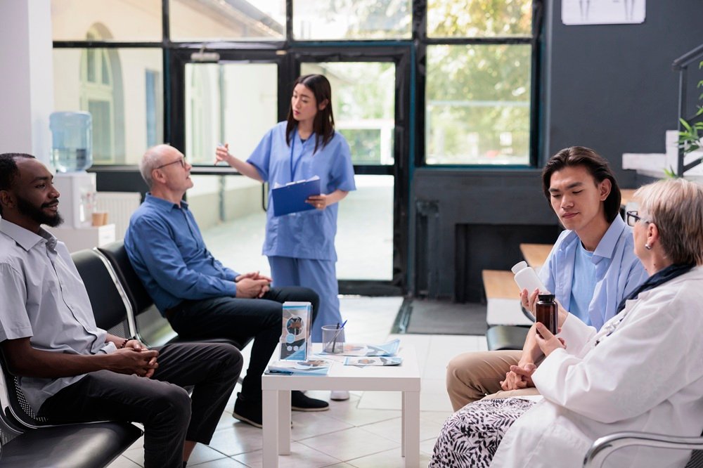 Elderly doctor sitting on chair in hospital waiting area holding antibiotic pills bottle while explaining medication treatment to asian patient during checkup visit consultation. Medicine service